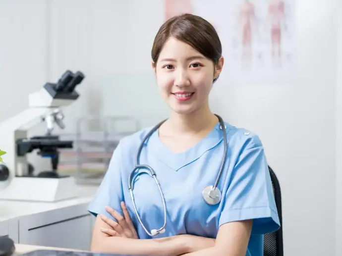 The background to this image is a a medical room of some sort.  There is a blurred poster on the wall of anatomical bodies to the right.  To the left, is a desk with a filing tray and next to it, a microscope, all also blurred out.  In the forefront is a young mediacl professional.  She has brown hair tied back off her face and is looking towards the camera and smiling.  She is wearing blue hospital scrubs and has a stehascope around her nexk.  She is sat down so we cannot see below her waiste level and she has her arms crossed across her body.